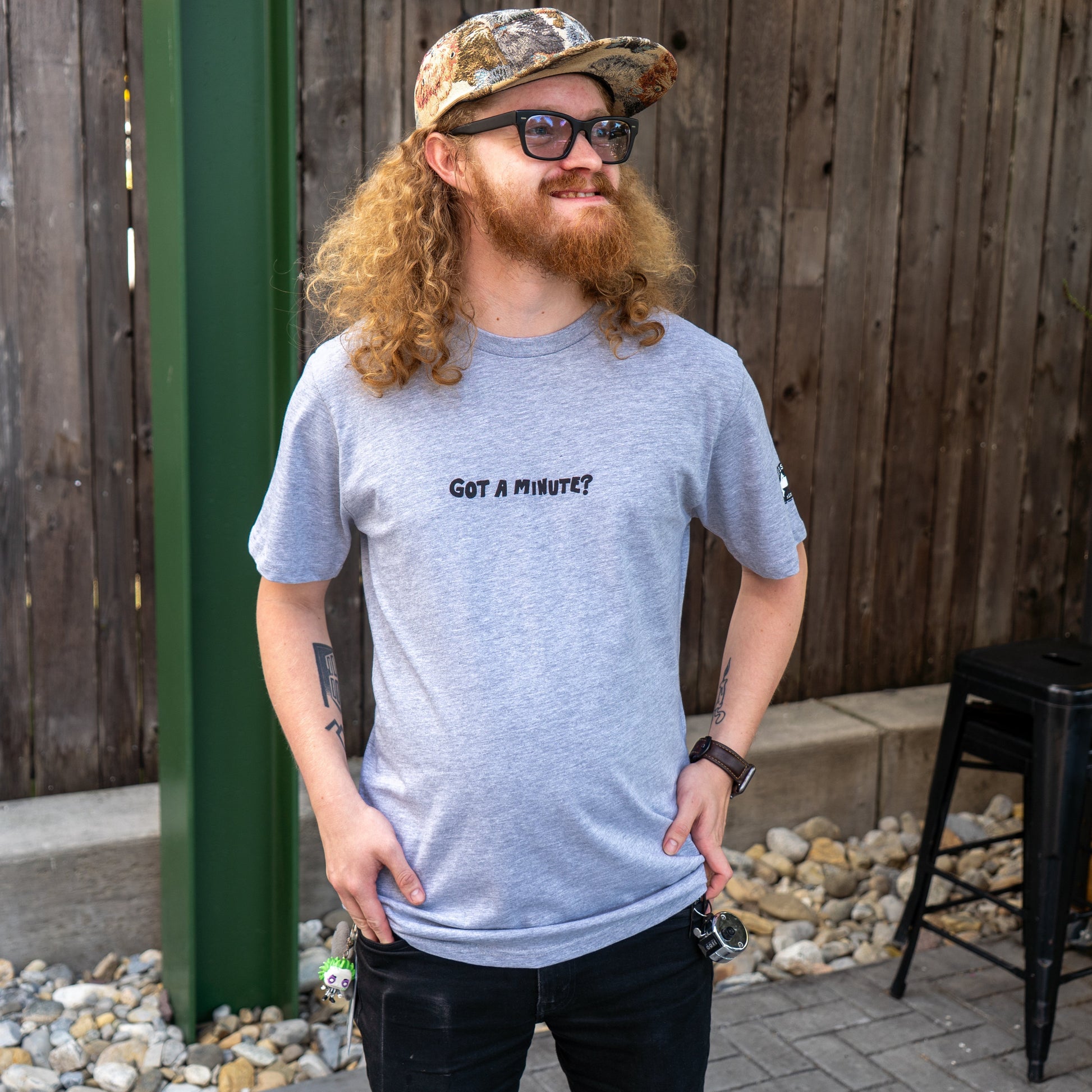 A smiling person with curly red hair and a beard stands outside by a wooden fence wearing sunglasses, a camouflage cap, and the GO WEST T-SHIRT COMPANY Grey Got A Minute Short Sleeve Tee with "GOT A MINUTE?" on the front. Their hand is in their pocket.