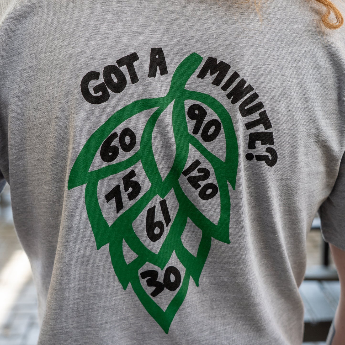 Someone wears the Grey Got A Minute Short Sleeve Tee by GO WEST T-SHIRT COMPANY, featuring a bold hop logo on the back with Minute Series numbers inside and "GOT A MINUTE?" in striking text above the graphic.