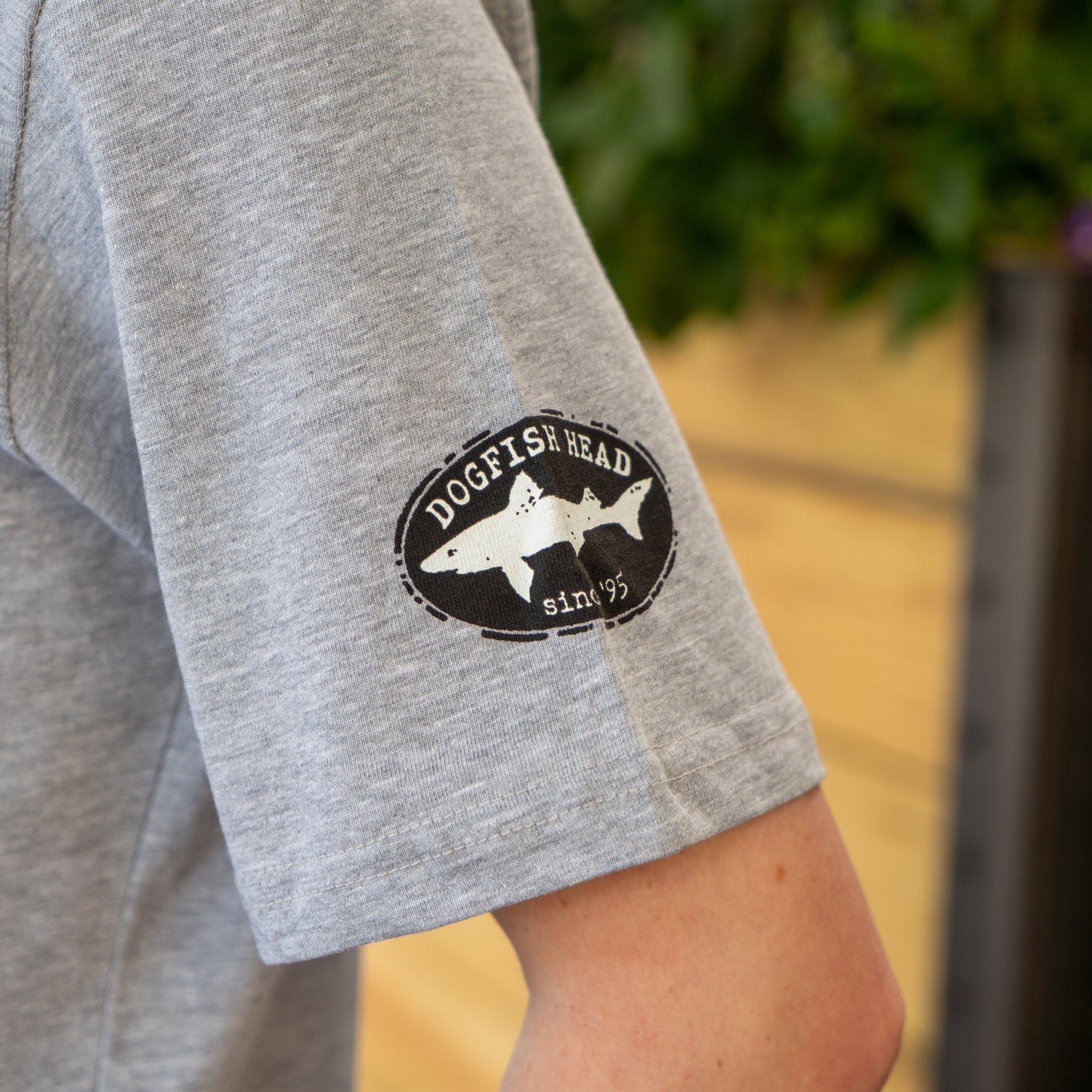 A close-up of a person's arm wearing the GO WEST T-SHIRT COMPANY Grey Got A Minute Short Sleeve Tee, featuring an oval Dogfish Head since '95 bold hop shark logo on the sleeve, with greenery and wood in the background.