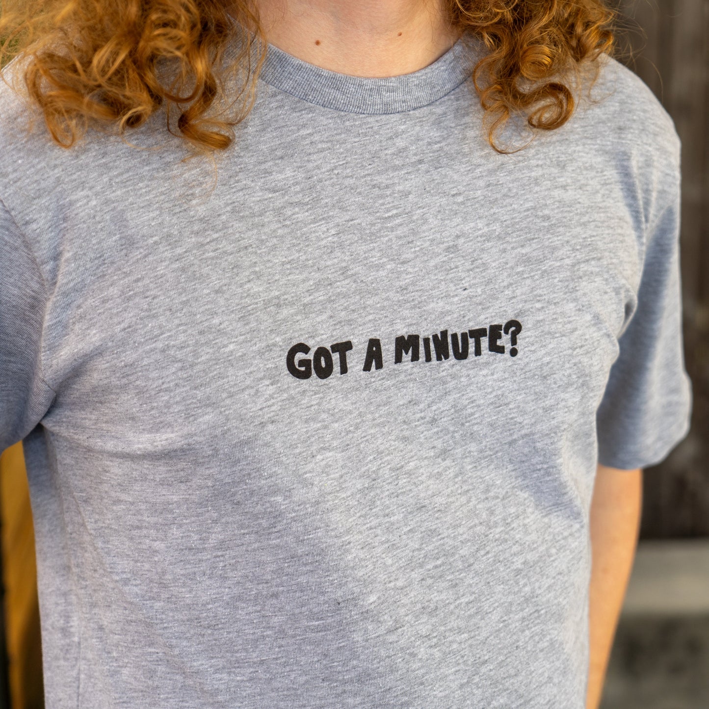 A person with long curly hair wears the GO WEST T-SHIRT COMPANY Grey Got A Minute Short Sleeve Tee, featuring a bold hop logo and “GOT A MINUTE?” in black on the chest. Their face is not visible in this Minute Series image.