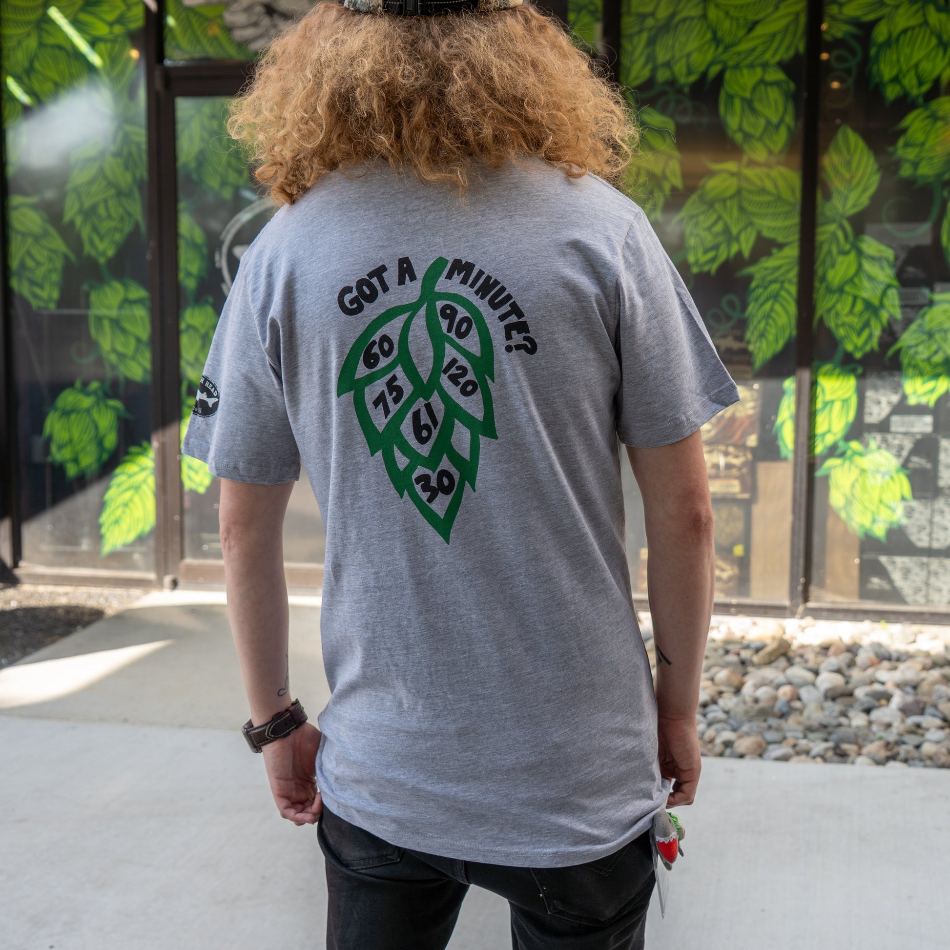 A person with curly hair wears the GO WEST T-SHIRT COMPANY's Grey Got A Minute Short Sleeve Tee, featuring a bold hop logo and “Got a minute? 60, 75, 90, 120, 6, 20” on the back—ideal for Minute Series fans—standing before a leafy mural.