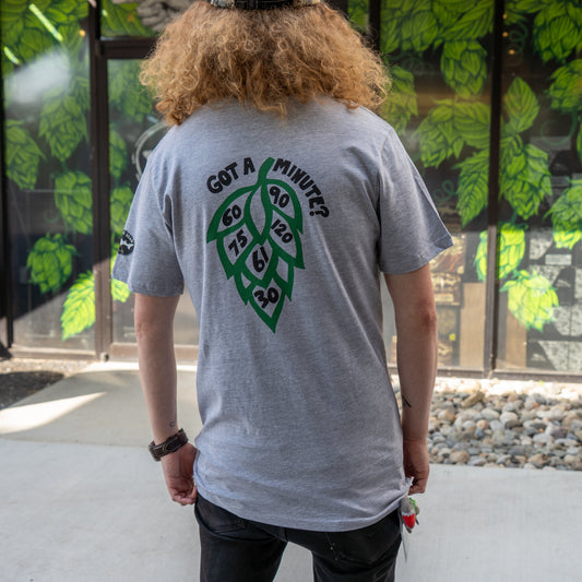 A person with curly hair wears the GO WEST T-SHIRT COMPANY's Grey Got A Minute Short Sleeve Tee, featuring a bold hop logo and “Got a minute? 60, 75, 90, 120, 6, 20” on the back—ideal for Minute Series fans—standing before a leafy mural.