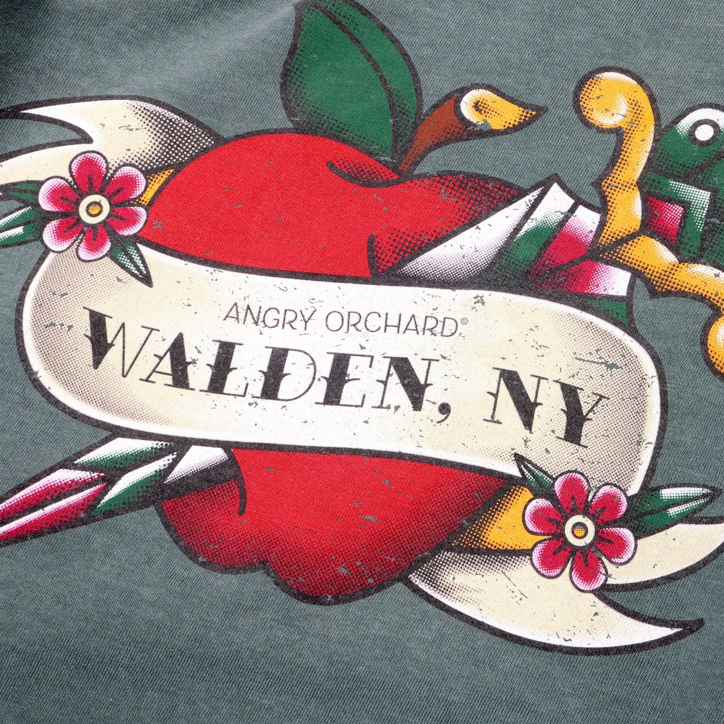 Close up of dark green long sleeve, with a design of a red apple being impaled by a dagger from top left shoulder to bottom right chest. The apple is wrapped in a banner and two small pink flowers. Banner reads "Angry Orchard, Walden, NY"