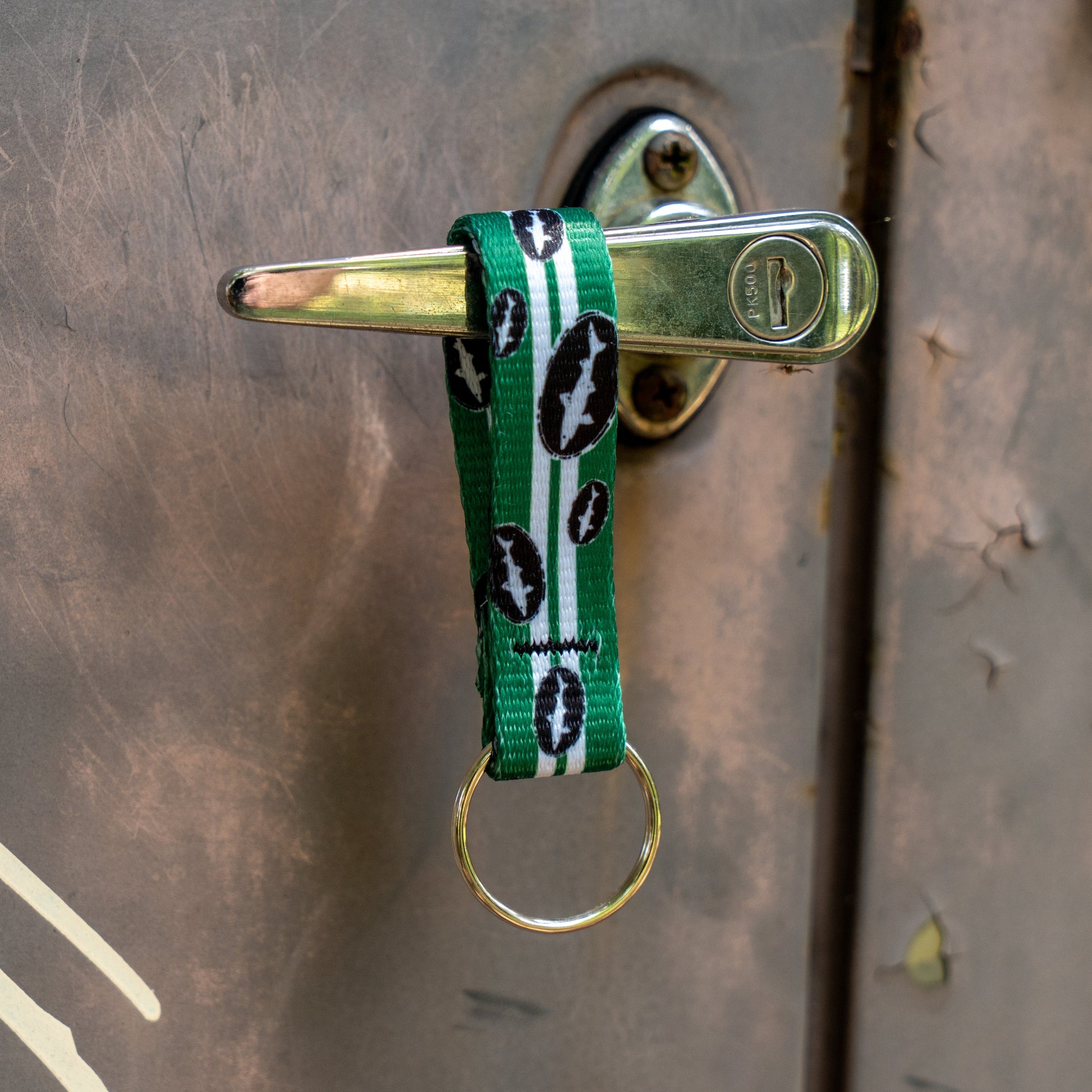 A Green Retro Keychain by Lupine, Inc. with black bat symbols hangs from a silver door handle. Its gold metal ring and green-and-white fabric add retro flair and good vibes to any entrance—ideal for designated drivers.