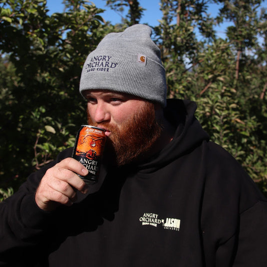 Man in Grey Carhartt Beanie with Angry Orchard logo reading "Angry Orchard, Hard Cider", drinking an angry orchard blood orange hard cider in a can