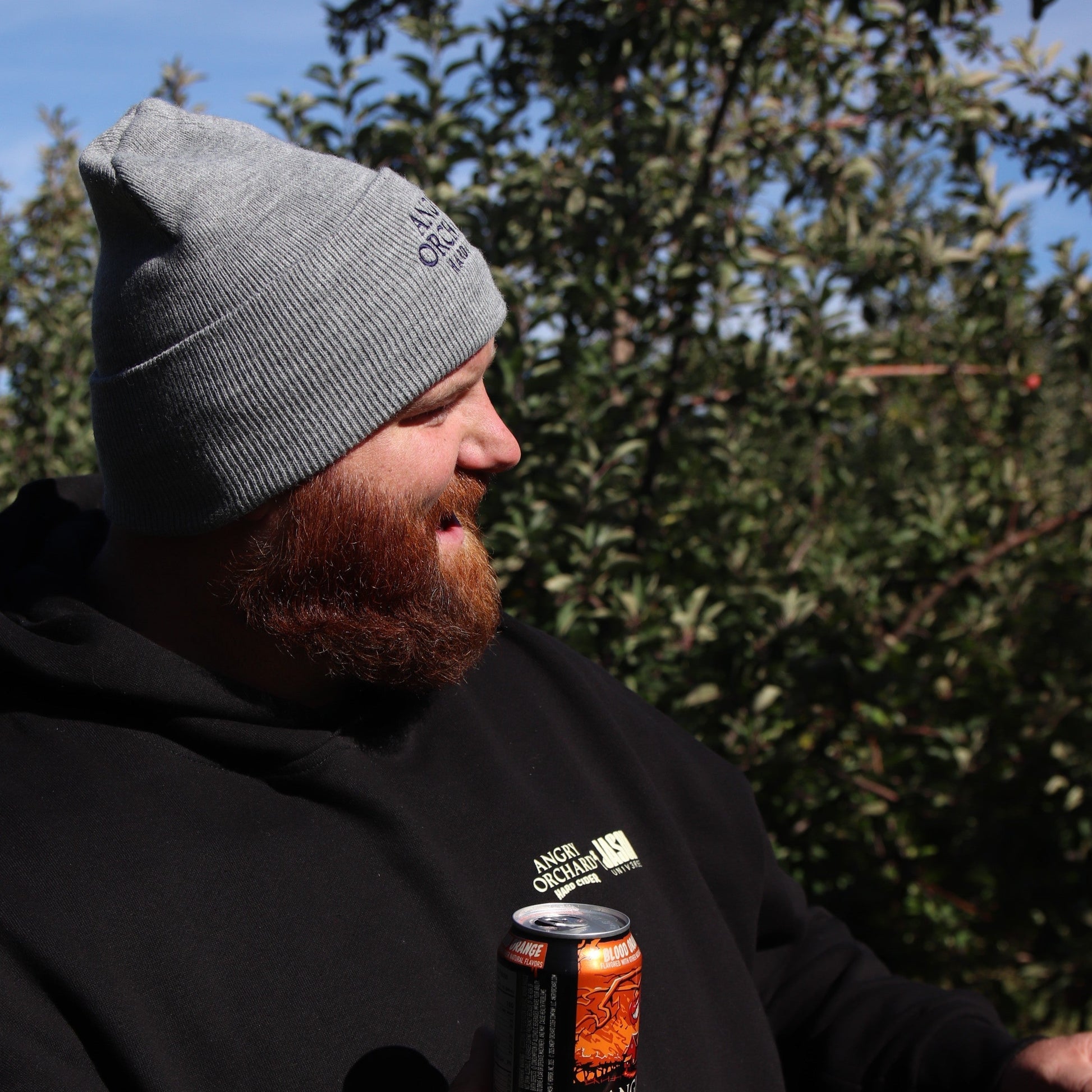 Man holding a blood orange angry orchard can outside, smiling wearing a grey Angry Orchard Hard Cider Carhartt Beanie