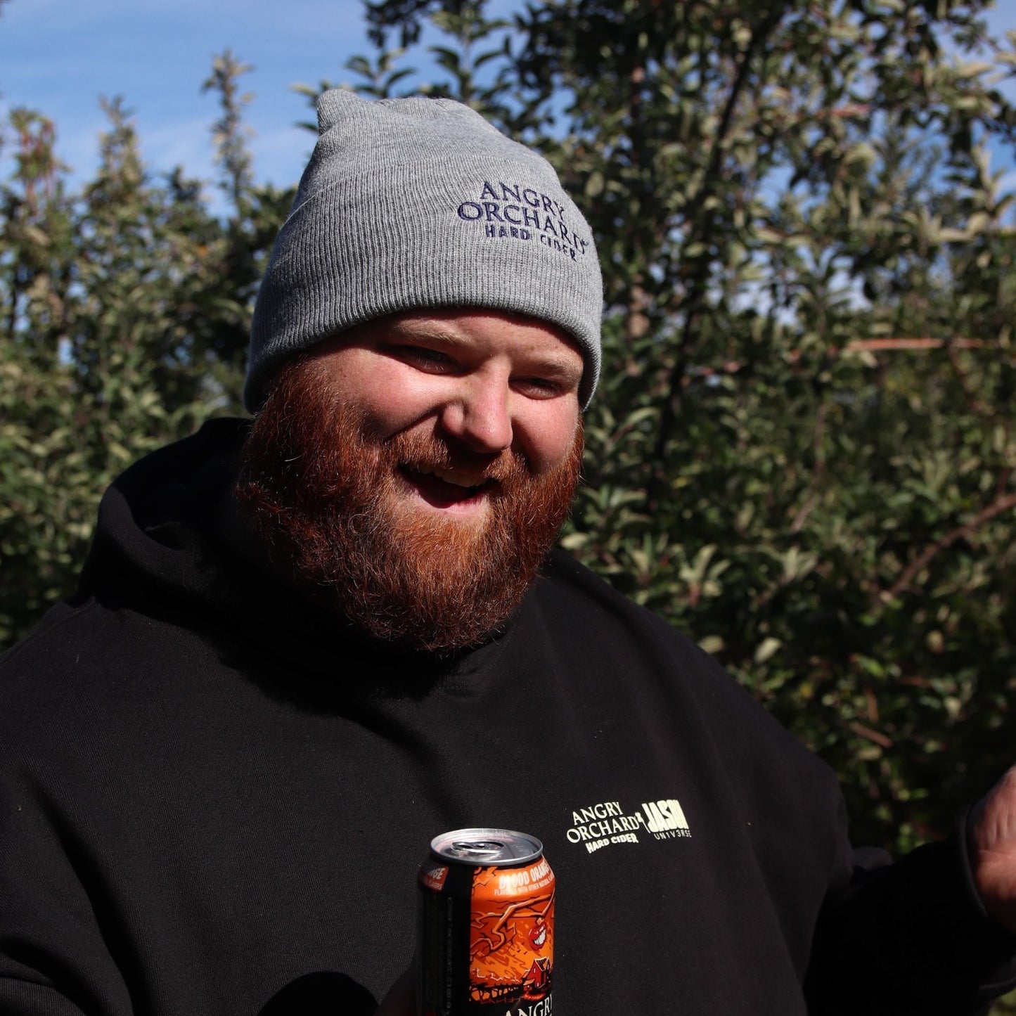 Man holding a blood orange angry orchard can outside, smiling wearing a grey Angry Orchard Hard Cider Carhartt Beanie