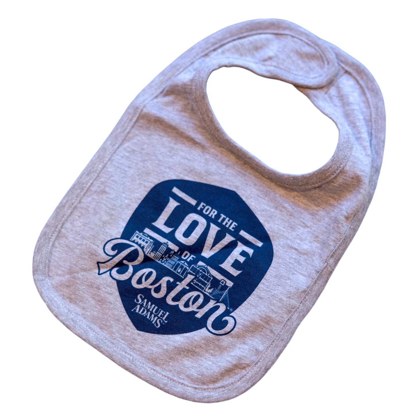 Grey bib with a blue shield in the center. Sheild features writing "For the Love of Boston" with the Sam Adams logo at the bottom of it. The Boston skyline is featured in between the words.
