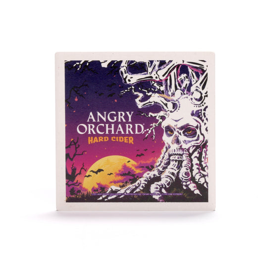 A square coaster with a Halloween-themed design. In the foreground, a gnarled tree with a skull-like face and boney branches stands against a purple and orange sky. A full yellow moon is visible in the background, with black bats flying across it. The text "ANGRY ORCHARD HARD CIDER" is written in white and orange letters to the left of the tree.
