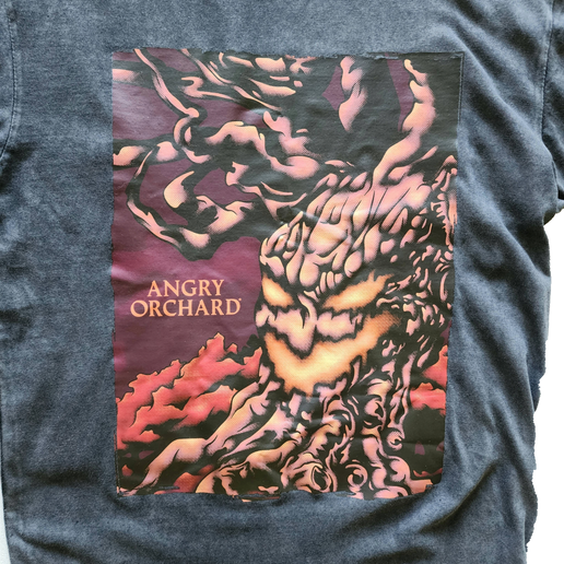 Close-up of front of black tee with image of "haunted tree" and writing "Angry Orchard". Tree is made to look like it has a face - like a jack-o-lantern. 