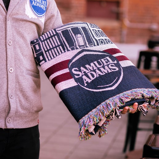 A 72" x 55" blanket that features the Boston skyline in white over a navy background and features the phrase "Here for the beer" in the center in white letters. Pictured here folded up to show only the Samuel Adams logo on the bottom middle.