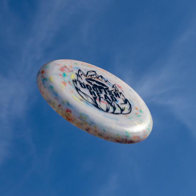 A white speckled Hop Design Frisbee by Arrasmith Promotions LLC, featuring a black logo, soars through the clear blue sky.