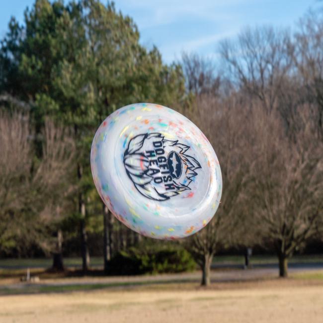 The Hop Design Frisbee from Arrasmith Promotions LLC, featuring a vibrant hop outline and Dogfish Head logo, flies through a park with green grass and tall trees in the background.