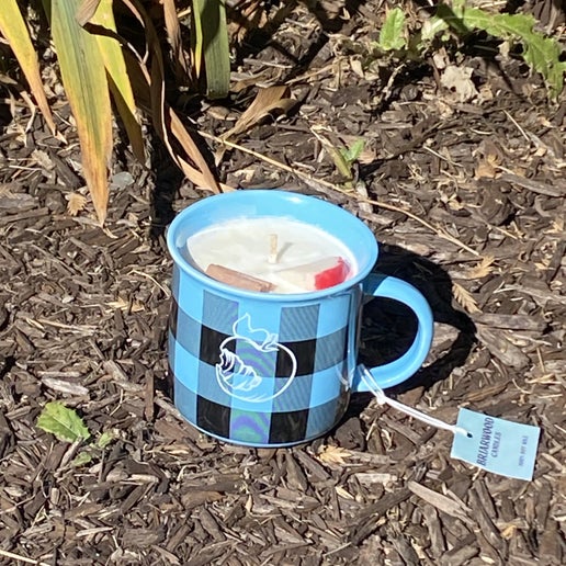 Backside of blue and black plaid mug with a candle in it. Backside of the mug in white is a logo of the Angry Orchard apple. The candle scent is Hot Apple Cider, containing a faux cinnamon stick and apple slice for decoration.