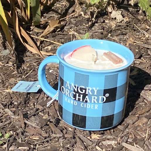Blue and black plaid mug with a candle in it sitting in mulch. On the front of the mug in white text it reads "Angry Orchard Hard Cider". The candle scent is Hot Apple Cider, containing a faux cinnamon stick and apple slice for decoration.