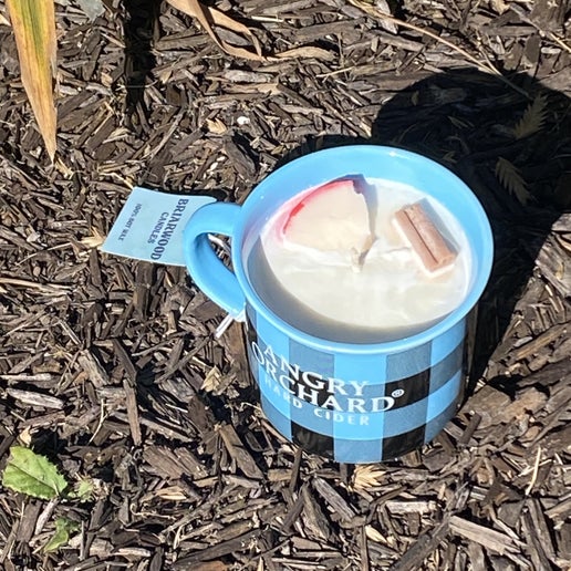 Blue and black plaid mug with a candle in it sitting in mulch. On the front of the mug in white text it reads "Angry Orchard Hard Cider". The candle scent is Hot Apple Cider, containing a faux cinnamon stick and apple slice for decoration.