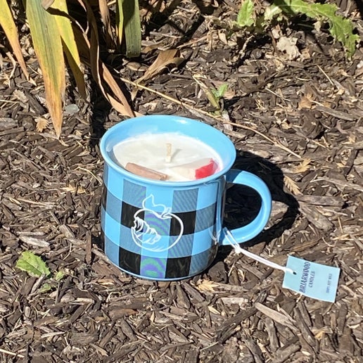 Backside of blue and black plaid mug with a candle in it. Backside of the mug in white is a logo of the Angry Orchard apple. The candle scent is Hot Apple Cider, containing a faux cinnamon stick and apple slice for decoration.