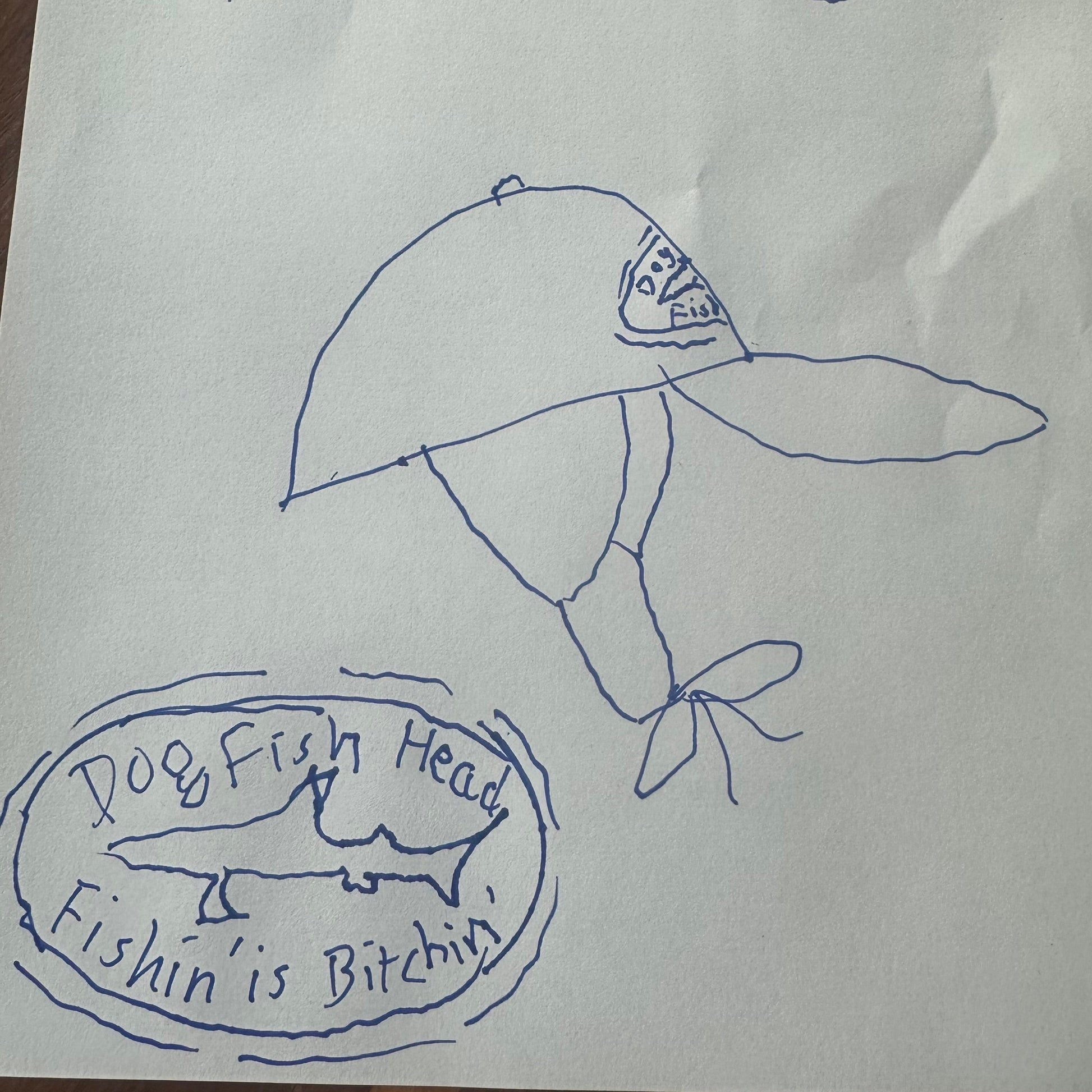 A hand-drawn blue pen illustration of the Fishin' is Bitchin' Retro Hat with Ear Flaps by Grandstand Glassware & Apparel, featuring a fish and the word "Fish" on the crown. Beneath, the text "Dogfish Head Fishin’ Is Bitchin’" appears alongside an oval design with a shark, reminiscent of the iconic Dogfish Head flap hat.