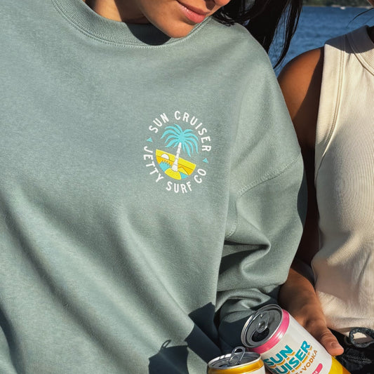 A person wearing a sage green crewneck sweatshirt with a circular 'Sun Cruiser Jetty Surf Co' graphic on the chest, holding a can of Mojave Yacht Soda outdoors near water.