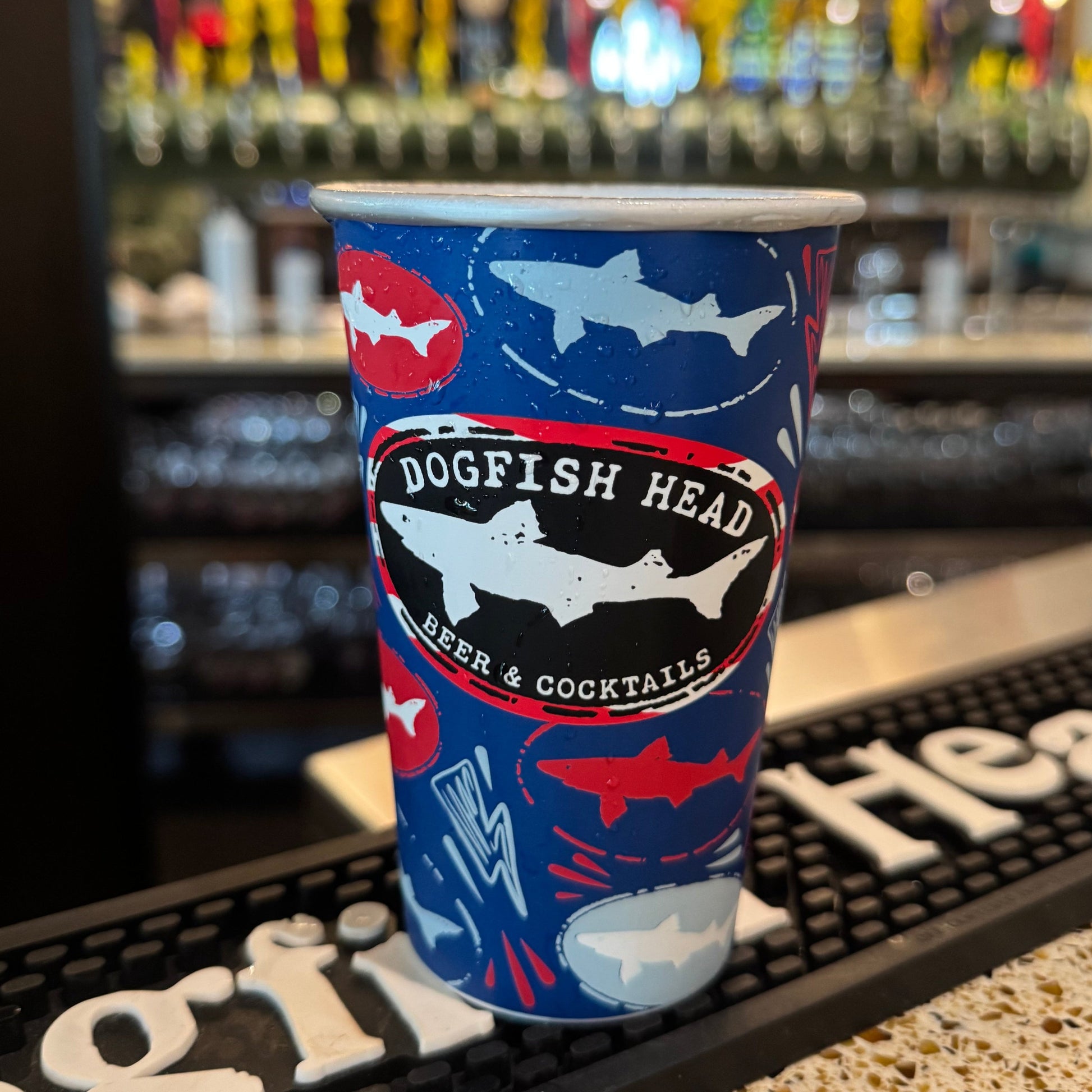 Dogfish Head aluminum can glass reusable cup with shark pattern sitting on bar mat, condensation visible, craft beer taproom background with blurred draft handles and menu board