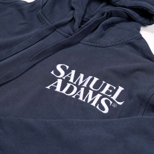 Indigo-colored hoodie with a front pocket and the classic Samuel Adams logo printed in white in the top left. Pictured close-up of the logo.