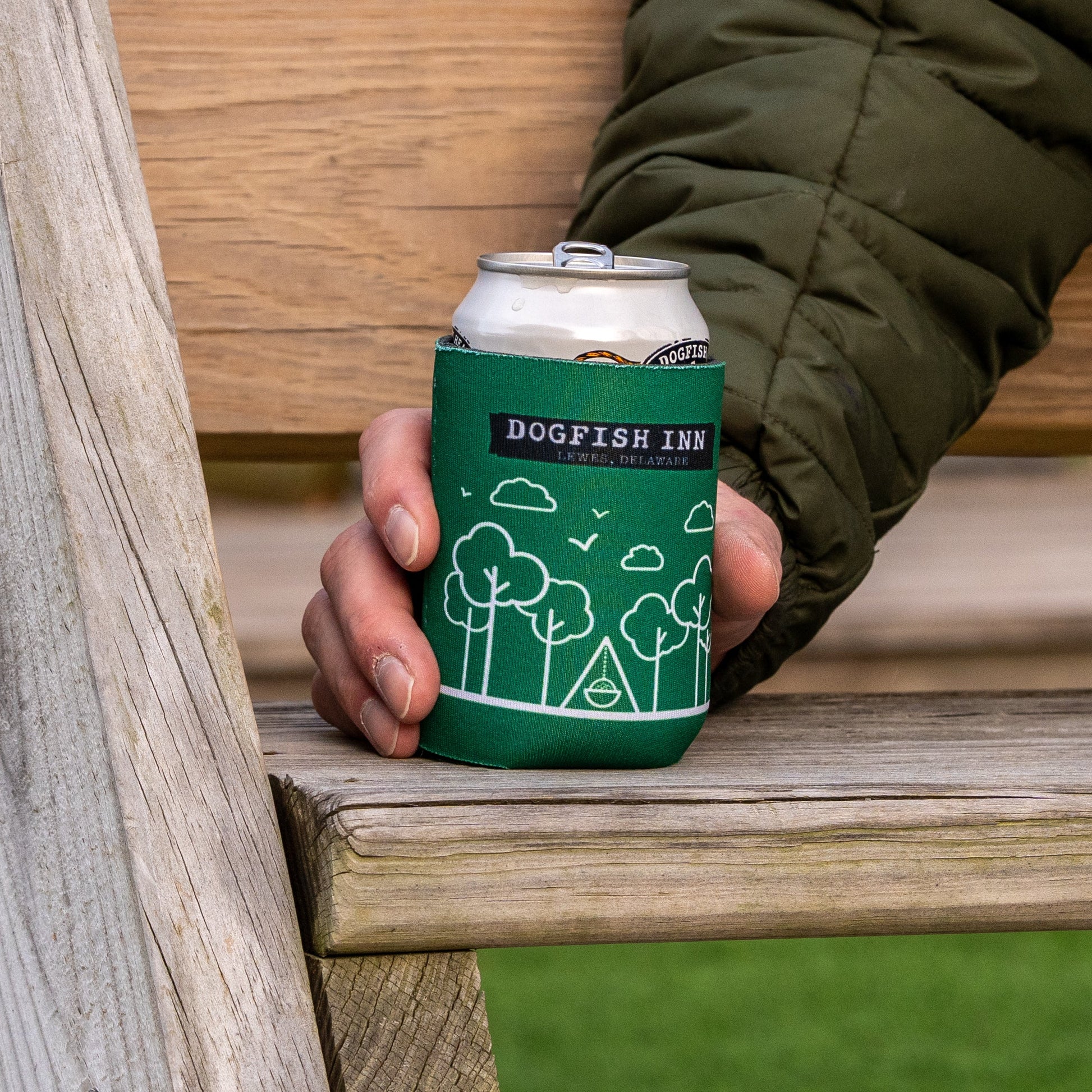 Someone in a green jacket holds a canned drink inside a Grandstand Glassware & Apparel Inn Logo Can Cooler with white tree and cloud designs, while sitting on a wooden bench.