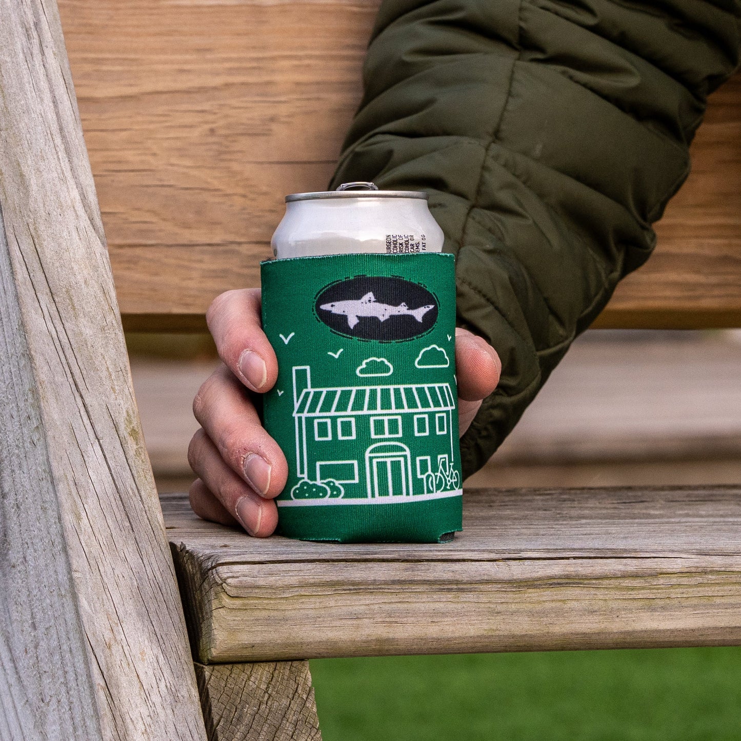 A person in a green jacket holds a can with the Inn Logo Can Cooler by Grandstand Glassware & Apparel, featuring a house design, while sitting on a wooden bench outdoors—ideal for fans of unique beer accessories.