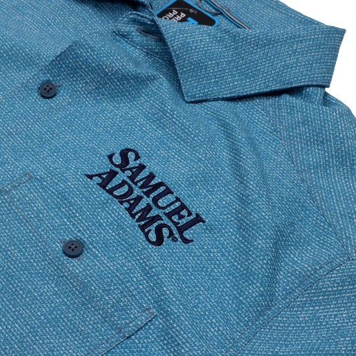 Front and back view of a light blue KUHL short sleeve button-up shirt featuring a textured pattern and Samuel Adams logo embroidered on the left chest.