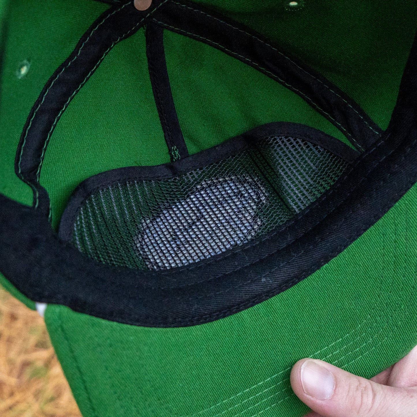 Close-up of the inside of a Merchtank Kelly Green Rope Hat, showing a white object in a black mesh pocket. A hand holds the brim of this one-size-fits-most cap.