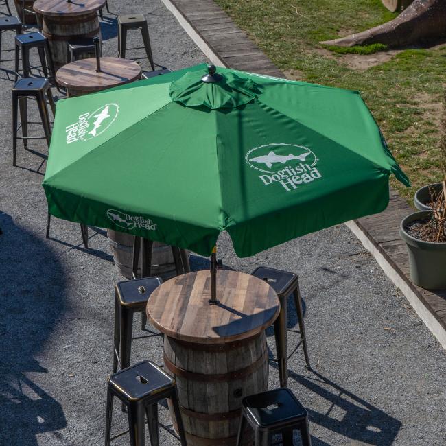 A Lg Logo Umbrella by Steamboat Marketing Inc shades a round wooden barrel table with barstools in an inviting gravel-covered outdoor seating area.