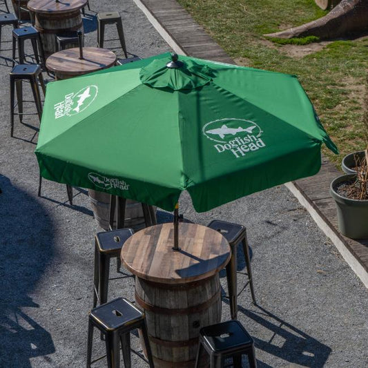 A Lg Logo Umbrella by Steamboat Marketing Inc shades a round wooden barrel table with barstools in an inviting gravel-covered outdoor seating area.