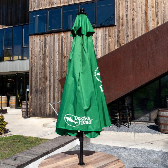 A closed green Lg Logo Umbrella by Steamboat Marketing Inc sits on a wooden table outside a modern building with wood paneling and large windows, ready to offer stylish sun protection when opened.