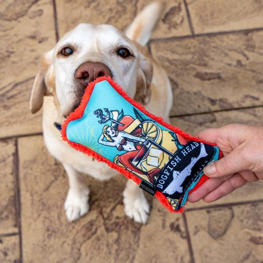 A yellow Labrador gazes up at the Cycle Dog LLC Mermaid Dog Toy, a colorful and durable bone-shaped toy featuring vibrant artwork, on a textured stone patio.