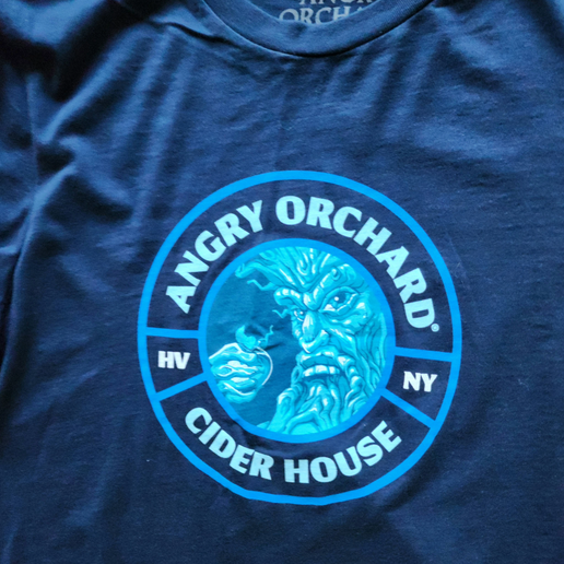 This is a navy tee with a light blue emblem centered to the tee. In the center is an image of the Angry Orchard tree trunk with a face on it. Around the image is writing including, "Angry Orchard Cider House", "HV", and "NY". The back of the tee is solid navy.