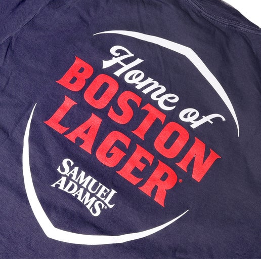 Backside close up of a navy shirt with a large Home of Boston Lager Samuel Adams shield shaped logo. Design is in white text with "Boston Lager" in red.
