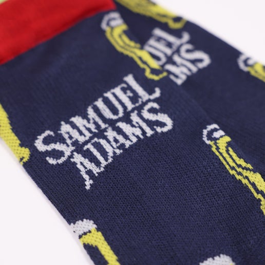 A pair of navy socks with red on the toe, heel, and top of ankle, and pints of beer with the Sam Adams logo on the ankle.