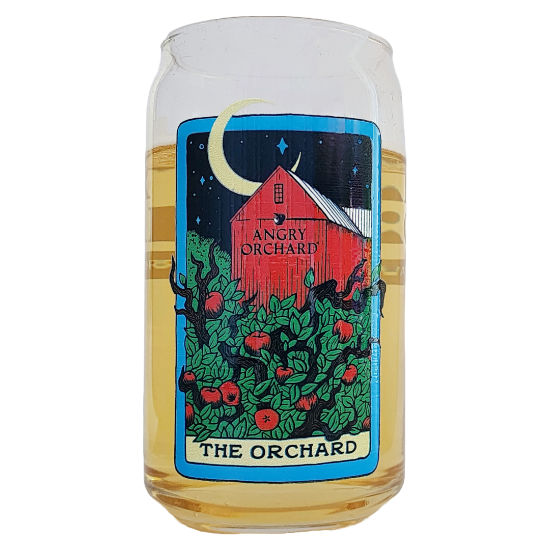 Angry Orchard glass filled with pale golden cider, featuring colorful illustrated artwork on the front. Reading like a tarot card, a bright red barn sits beneath a dark blue night sky with a yellow crescent moon, surrounded by deep green apple trees dotted with red apples, with a cream colored label reading "The Orchard" at the bottom.