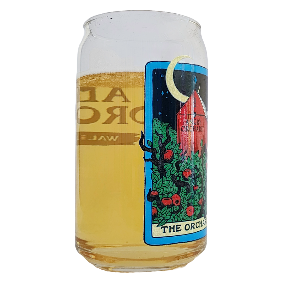 Side angle view of an Angry Orchard cider glass showing golden cider inside. The illustrated orchard label wraps around the glass with green leafy branches and red apples.