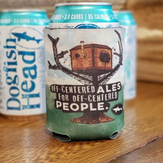 A 12oz Dogfish Head beer is kept cool in the OCA Treehouse Can Cooler by Grandstand Glassware & Apparel, showcasing a quirky treehouse design and “Off-centered ales for off-centered people.” Two standard cans are blurred in the background.