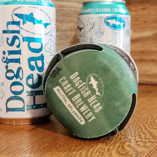 Two OCA Treehouse Can Coolers from Grandstand Glassware & Apparel rest on a wooden table, with one cooler lying on its side to display the logo and text stamped on the bottom.