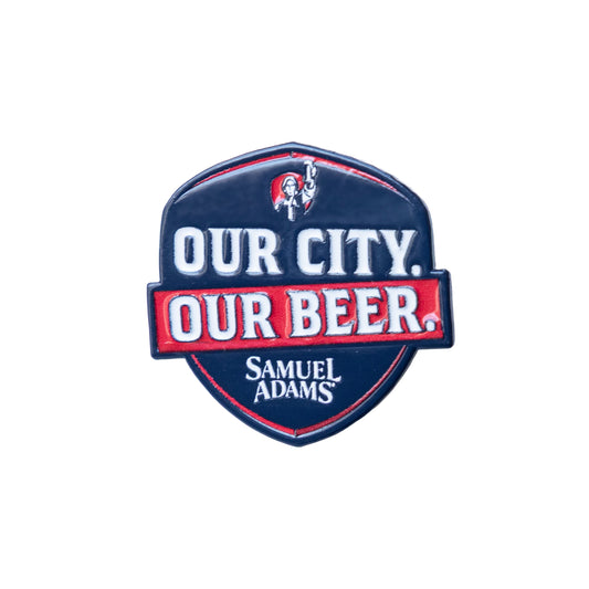 Samuel Adams enamel pin in shield shape featuring navy blue background with white “OUR CITY.” text, red banner reading “OUR BEER.” in white lettering, red outline accents, small Samuel Adams revolutionary figure icon at the top, and “Samuel Adams” text at the bottom