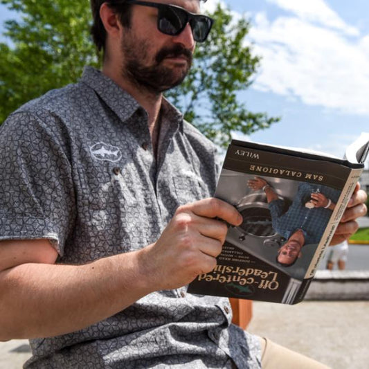A man in sunglasses and a patterned short-sleeve shirt sits outside reading Book- Off-Cent Leadership by J Wiley and Sons Publishing, surrounded by trees and a blue sky.