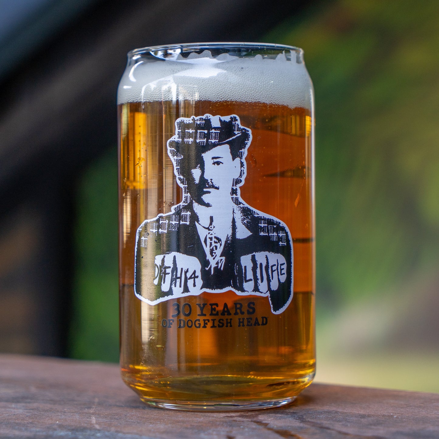 An Off Centered Way Can Glass by Grandstand Glassware & Apparel, filled with amber beer and topped with foam, sits on wood. The glass displays a black-and-white man in vintage attire and "30 Years of Dogfish Head" text.