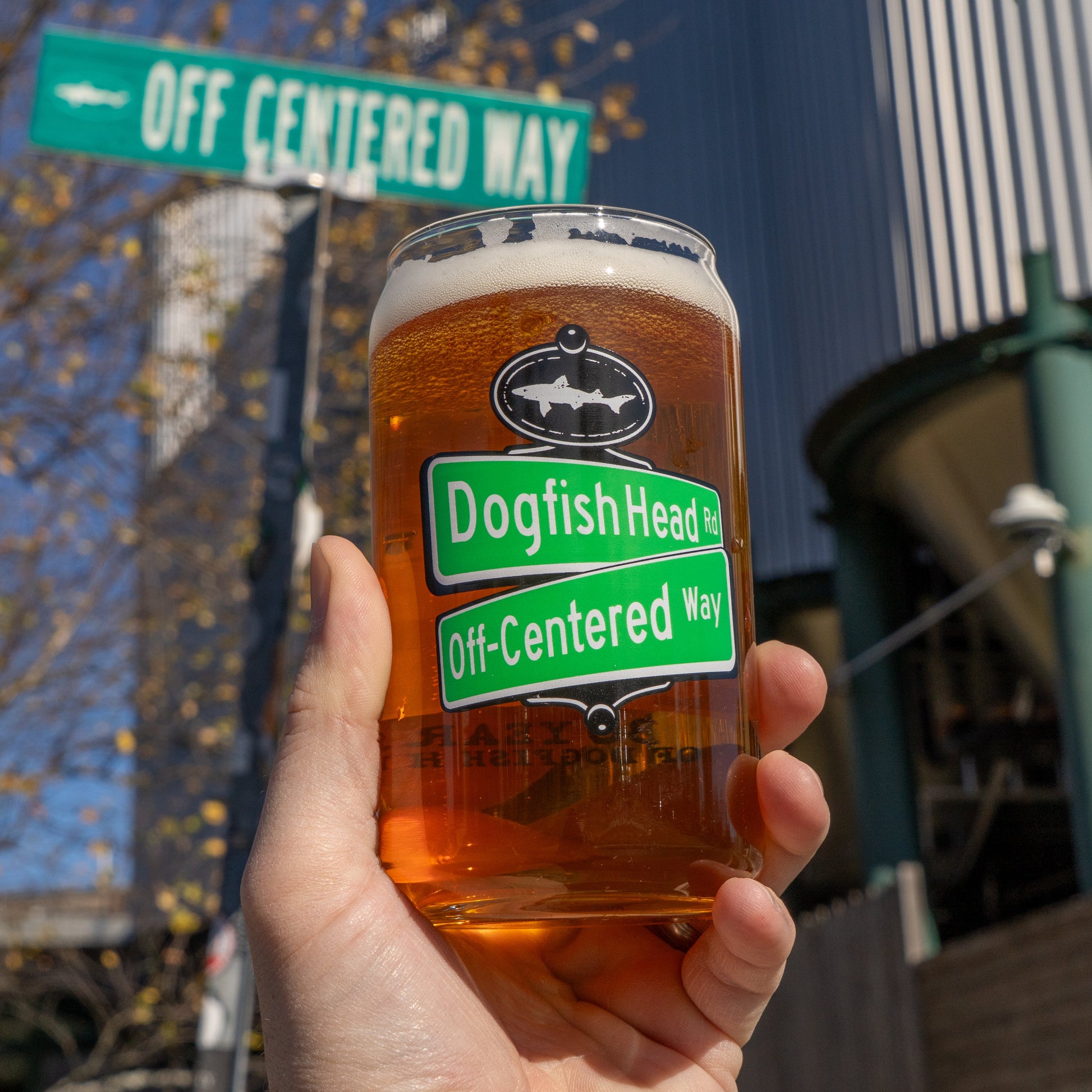 A hand holds a Grandstand Glassware & Apparel Off Centered Way Can Glass filled with amber beer, its label matching a street sign—both celebrating 30 years—set before a modern building and clear blue sky.