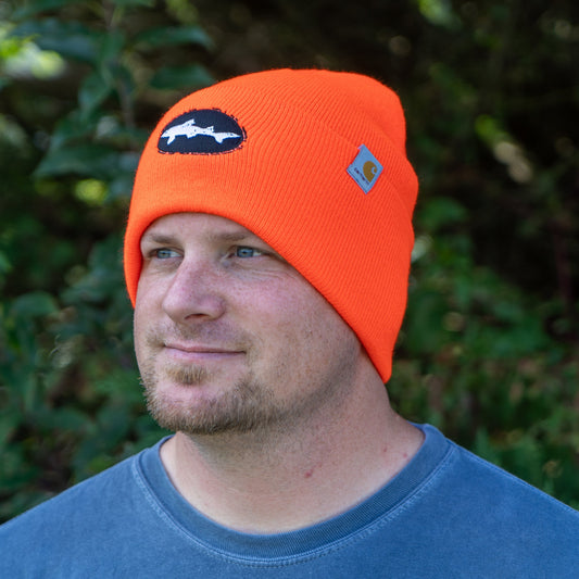A man with a short beard stands outdoors amid greenery, wearing a warm, bright Orange Beanie by Mirror Image Inc.