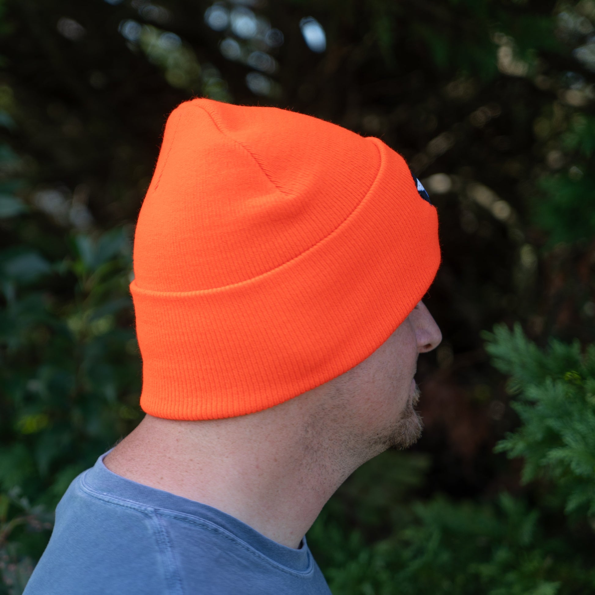 A person wearing the Orange Beanie by Mirror Image Inc. and a blue-gray Carhartt shirt is seen in profile, looking away from the camera, with green foliage in the background.