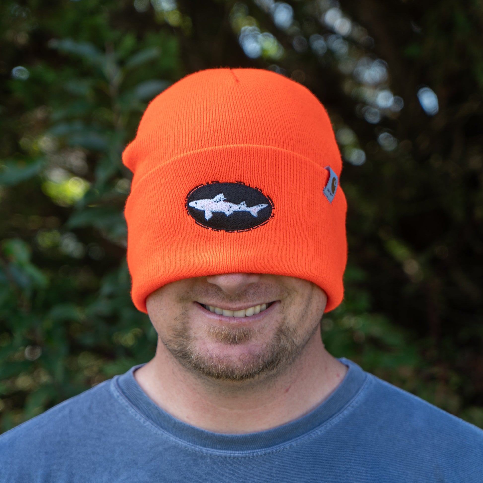 A person smiles warmly outdoors, wearing a bright orange Mirror Image Inc. Orange Beanie pulled low over their eyes, featuring a shark patch on the front, with greenery in the background.