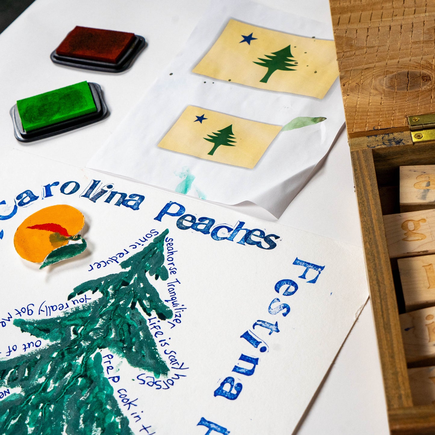 Close-up of Festina Peche screen print poster in progress with green and orange inks, rubber stamps, and carved print blocks on a studio work surface