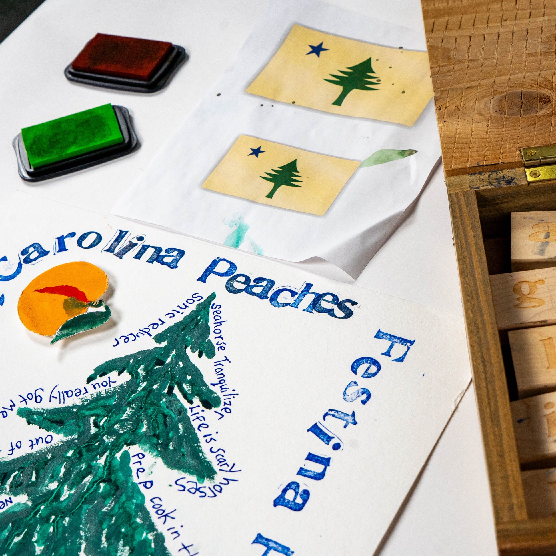 Close-up of Festina Peche screen print poster in progress with green and orange inks, rubber stamps, and carved print blocks on a studio work surface
