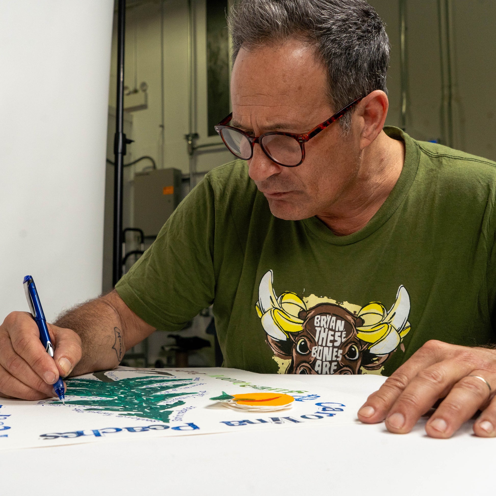 Artist hand-lettering details on Festina Peche screen print poster at a printmaking table, surrounded by ink pads and tools in a studio setting.
